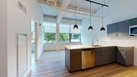 This single-floor unit in Hartford, CT showcases a compelling blend of industrial and contemporary design. Soaring ceilings with exposed structural beams, ductwork, and track lighting lend a loft-like character throughout the open living space. Light wood-tone flooring flows seamlessly across the main area, enhancing the sense of openness and warmth.

The kitchen is a standout feature, offered in configurations that include a center island or an L-shaped layout. Dark gray cabinetry pairs with light countertops and a full suite of stainless steel appliances, including a refrigerator, dishwasher, and microwave. Matte black pendant lights hang above the island and counters, adding a refined industrial accent. Large windows fitted with roller shades allow generous natural light and views of surrounding greenery.

A bathroom features clean white walls, large-format neutral tile flooring, a full tub and shower, and a dark-framed glass door entry. A dedicated closet with built-in shelving provides practical storage. The overall ambiance is modern and airy, combining functional design with architectural character well-suited to urban living in Hartford.