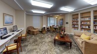 This single-floor community library and lounge is a thoughtfully designed space that blends functionality with a warm, inviting ambiance. The room is anchored by floor-to-ceiling built-in bookshelves lining multiple walls, housing an extensive collection of books that cater to a wide range of reading interests. Bright flush-mounted ceiling lights illuminate the space evenly throughout, creating a comfortable environment for reading and socializing.

A cozy seating area at the heart of the room features a plush sofa accented with a yellow throw pillow, arranged around a classic wooden coffee table adorned with floral décor. Patterned carpeting with a subtle botanical motif adds warmth and visual interest underfoot throughout the entire space.

A dedicated computer workstation area offers two desktop terminals set along a built-in desk surface, flanked by additional bookshelves and framed nautical artwork, making it an ideal spot for research or correspondence.

One of the room's most distinctive architectural features is an elegantly arched alcove that frames a circular dining nook, complete with a round table and colorful upholstered chairs. Large windows with adjustable blinds allow natural light to filter in, while a pendant light fixture adds a refined finishing touch. A set of glass-paned French doors connects seamlessly to an adjacent area, enhancing the open, communal feel of this well-appointed gathering space.