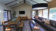 This single-floor space makes a striking first impression with its soaring vaulted ceiling accented by dark exposed wooden beams and recessed lighting, creating a sense of openness and architectural character. The living area is generously proportioned, anchored by dark hardwood flooring that runs throughout and complements the warm, earthy tones of the interior design. A wall-mounted flat-screen television is positioned above a rustic wooden media console, serving as a natural focal point in the room. Lush indoor plants add a vibrant, organic element to the space, softening the clean lines of the architecture. Comfortable dark leather sofas are arranged to encourage conversation and relaxation, accented with coral and neutral throw pillows that introduce a pop of color. A large, dark drum pendant light fixture overhead adds a bold, modern design statement. Natural light pours in generously through large windows and glass doors along one wall, offering views to an outdoor area and keeping the interior bright and inviting. A rustic farmhouse-style dining table with matching benches provides a casual dining or gathering area, reinforcing the space's modern rustic aesthetic throughout.