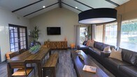 This single-floor space makes a striking first impression with its soaring vaulted ceiling accented by dark exposed wooden beams and recessed lighting, creating a sense of openness and architectural character. The living area is generously proportioned, anchored by dark hardwood flooring that runs throughout and complements the warm, earthy tones of the interior design. A wall-mounted flat-screen television is positioned above a rustic wooden media console, serving as a natural focal point in the room. Lush indoor plants add a vibrant, organic element to the space, softening the clean lines of the architecture. Comfortable dark leather sofas are arranged to encourage conversation and relaxation, accented with coral and neutral throw pillows that introduce a pop of color. A large, dark drum pendant light fixture overhead adds a bold, modern design statement. Natural light pours in generously through large windows and glass doors along one wall, offering views to an outdoor area and keeping the interior bright and inviting. A rustic farmhouse-style dining table with matching benches provides a casual dining or gathering area, reinforcing the space's modern rustic aesthetic throughout.