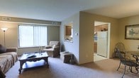 This single-floor residential space offers a functional and comfortable layout designed to maximize openness and ease of living. The living area is finished with wall-to-wall carpet in a neutral tone, creating a warm and inviting atmosphere. A large window dressed with vertical blinds allows natural light to fill the room, enhancing the sense of space. The living area connects seamlessly to the kitchen through a pass-through window, providing a convenient point of interaction between the two spaces. The kitchen is well-appointed with medium-toned wood cabinetry, a white refrigerator, and a gas or electric range, offering ample storage and workspace for everyday use. The light-colored tile flooring in the kitchen contrasts with the carpeted living space, clearly delineating the two functional areas. Neutral wall tones throughout the space contribute to a clean and cohesive aesthetic. The overall layout is efficient and practical, making thoughtful use of the available floor space. The open sightlines between the living and kitchen areas lend the space an airy, connected feel well-suited for everyday living.