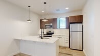 This thoughtfully designed single-floor unit offers a clean, modern aesthetic throughout its well-utilized footprint. Light-toned hardwood flooring runs continuously through all areas, creating a cohesive and airy feel.

The kitchen is a standout feature, showcasing a large waterfall quartz island with an undermount sink and matte black faucet, illuminated by sleek pendant lighting. Two-toned cabinetry combines white lower cabinets with dark walnut upper cabinets, complemented by a subway tile backsplash. Stainless steel appliances — including a range, over-the-range microwave, and refrigerator — complete the functional and stylish cooking space.

The bedroom offers a comfortable and uncluttered retreat, featuring a ceiling fan, a walk-in closet, and a window that allows natural light to fill the room. The clean white walls and neutral tones give the space a calm, versatile ambiance suitable for a variety of furnishing styles.

A hallway connects the primary areas and includes recessed lighting and a utility panel, adding to the unit's practical design. Windows positioned throughout the space invite natural light while maintaining privacy. The overall design reflects a contemporary sensibility with quality finishes, making this a well-appointed and efficiently organized living space.
