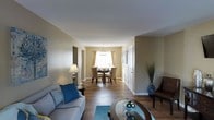 This single-floor residence offers a thoughtfully arranged open-concept layout that connects the living and dining areas in a seamless flow. The living room serves as the centerpiece of the space, featuring rich, dark hardwood floors that run throughout and warm beige walls that create an inviting, neutral backdrop. A light gray sofa accented with blue and yellow throw pillows anchors the seating area, complemented by a round glass-top coffee table and a teal area rug that adds a pop of color underfoot. A leather accent chair and floor lamp provide additional comfort and ambiance.

French doors at the far end of the living room flood the space with natural light while offering a view to the outdoors, enhancing the open and airy feel of the room. The smooth, light-toned ceiling contributes to the sense of spaciousness.

The adjoining dining area is visible from the living room, centered around a round dark wood table surrounded by upholstered dining chairs. A delicate chandelier overhead adds an elegant touch, while a window dressed with sheer gold curtains provides soft, natural light. The cohesive flooring and color palette throughout create a harmonious and well-balanced living environment.