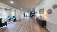 This well-appointed single-floor space offers a seamlessly connected open layout designed for comfort and functionality. Wide-plank hardwood-style flooring flows continuously throughout, tying each area together in a cohesive, inviting aesthetic.

The expansive main hall greets visitors with recessed lighting, large windows dressed in sheer drapes, and double French doors that invite natural light. A sitting nook features a pair of teal wingback chairs, adding a bold accent to the otherwise neutral palette.

The living area serves as a central gathering point, anchored by a striking teal-paneled fireplace wall with a mounted television above. Crown molding and recessed lighting add refinement, while large windows and French doors open the space visually to the outdoors.

Adjacent to the living area, a casual dining zone features high-top tables and bar-height seating, accentuated by a large-scale Highland cattle canvas print. Windows along the wall fill the space with natural light.

The kitchen is both spacious and well-equipped, showcasing sage-green cabinetry with cross-panel detailing, light granite countertops, stainless steel appliances, and track lighting. A generous wraparound counter provides ample prep and serving space, with an open pass-through connecting seamlessly to the living and dining areas.