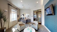 This single-level business center offers a thoughtfully designed workspace that balances functionality with a welcoming aesthetic. The main area features a dedicated workstation zone equipped with dual computer monitors and ergonomic seating at a sleek, dark-toned desk, making it well-suited for focused tasks or remote work needs. Framed artwork adorns the neutral-toned walls, adding a polished touch to the professional environment.

A lounge area in the foreground includes a pair of striped upholstered armchairs arranged around a round white accent table, providing a comfortable spot for informal meetings or a brief respite. Decorative teal glass vases on the table introduce subtle color accents that complement the room's soft, neutral palette.

Natural light streams in through a large arched window fitted with curtains, brightening the space alongside recessed ceiling lighting and a pair of elegant gold pendant lights that anchor the central area. A glass-paneled door leads to an adjacent private room, visible in the background, extending the overall utility of the center. The combination of warm textures, modern furnishings, and thoughtful lighting creates a productive yet inviting atmosphere.