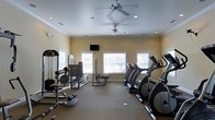 Located at 111 Coopers Ridge Blvd in Ladson, South Carolina, this fitness center offers a well-equipped, single-floor workout facility designed to meet a variety of exercise needs. The space is finished with warm yellow walls and white crown molding, giving it a clean and inviting atmosphere. Recessed lighting is complemented by dual ceiling fans, ensuring the room remains bright and well-ventilated throughout any workout session. Multiple windows along the exterior wall allow natural light to fill the space, creating an open and airy environment. The cardio section is well-stocked with a row of treadmills and elliptical machines, ideal for sustained aerobic exercise. Strength training is supported by a selection of cable and resistance machines, providing users with options for a comprehensive full-body workout. A wall-mounted television is positioned for easy viewing during cardio sessions, adding an entertainment element to the experience. The carpeted flooring provides comfort underfoot and contributes to a quieter exercise environment. Overall, this fitness center combines functional equipment with a thoughtfully designed space, making it a practical and comfortable amenity for residents and visitors of Coopers Ridge.