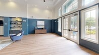 Located at 9198 Integra Hills Ln in Ooltewah, Tennessee, this generously sized single-floor community clubhouse offers a well-appointed, multi-functional layout designed for gathering and recreation. The main living area immediately impresses with soaring ceilings, bold navy-blue wainscoting, and light hardwood flooring that flows throughout the space. Oversized windows and French doors flood the room with natural light while offering views of the outdoor pool area. A striking built-in wet bar with patterned tile backsplash and charcoal cabinetry serves the main lounge, which also features a full-size billiards table beneath a statement chandelier. An adjacent seating area is thoughtfully arranged with contemporary gray armchairs, a marble-top coffee table, and curated blue wall art accented by a sliding barn door. The kitchen and living combination area showcases dramatic white quartz countertops with gold veining, dark cabinetry, stainless steel appliances, and a wall-mounted television. A dedicated game room with patterned carpet, ping-pong table, and natural light from multiple windows provides additional recreational space. Flexible open rooms with clean white walls and light wood floors offer versatile space for programming or events, completing this well-designed community facility.