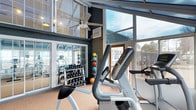 This fitness center is designed to provide an energizing and well-lit workout environment. The space is defined by expansive floor-to-ceiling windows that flood the interior with natural light and offer open views of the surrounding outdoors, creating an airy, motivating atmosphere. The soaring ceiling height adds a sense of spaciousness, and a ceiling fan overhead helps maintain comfortable airflow throughout the room.

The facility is thoughtfully equipped to support a variety of workout styles. Cardio machines, including ellipticals and a stationary bike, are positioned to take full advantage of the natural light and outdoor views. A dedicated free weights area features a well-organized dumbbell rack spanning a broad range of sizes, reflected in a large wall mirror that enhances the sense of depth within the space. A medicine ball rack adds to the strength training options available.

The neutral-toned carpet underfoot provides comfort during workouts, while the dark-painted walls and structural framing lend the space a modern, purposeful aesthetic. The combination of abundant natural light, high ceilings, and comprehensive equipment makes this fitness center a functional and inviting space for daily exercise.