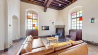 Located at 1006 W Lake St in Minneapolis, Minnesota, this remarkable single-floor space is a beautifully preserved historic structure that blends old-world architectural grandeur with modern furnishings. The interior is defined by soaring vaulted and beamed ceilings, arched doorways and windows, and warm terracotta tile flooring laid in a classic checkerboard pattern throughout.

The space is illuminated by an array of tall, multi-paned stained glass windows that cast vibrant hues of red, yellow, blue, and orange across the interior, creating a dynamic and ever-changing atmosphere. Two ornate stone fireplaces serve as dramatic focal points — one featuring an intricately carved stone surround with sculptural reliefs and herringbone brick interior, and another with a similarly decorative mantle.

The main gathering area is furnished with a generous leather sectional sofa, upholstered accent chairs, and high-top bar tables with modern seating, making the space well-suited for both relaxed lounging and social gatherings. Decorative wrought iron lantern sconces and chandelier pendants add warmth and historic character to the whitewashed plaster walls. Arched passageways connect distinct areas of the floor, offering both openness and intimate alcoves within this truly one-of-a-kind Minneapolis space.