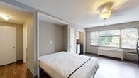 Located at 516 W Ormsby Ave in Louisville, Kentucky, this single-floor studio unit offers a compact and functional layout designed to maximize its available space. The main living area features durable hardwood-style flooring in a warm wood tone, neutral gray walls, and a decorative ceiling fan with a light fixture that provides ambient illumination throughout the room. A murphy bed folds out from a built-in wall unit, offering a space-saving sleeping solution without compromising the open feel of the living area. Natural light enters through a window fitted with blinds, overlooking greenery outside, and a wall-mounted HVAC unit provides climate control.

A compact kitchen alcove is tucked efficiently into the space, equipped with white upper cabinetry, a black gas range, and a sink. A full-size black refrigerator stands adjacent to the main living area, adding practical storage. Additional doorways lead to separate areas within the unit, including a bathroom featuring classic white subway-style wall tiles surrounding a full bathtub, with a window providing natural light and ventilation. The overall layout is straightforward and well-organized, making practical use of the single-floor footprint while maintaining a clean, neutral aesthetic throughout.