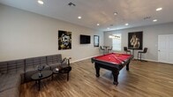 This clubhouse is a well-appointed single-floor community gathering space designed for recreation and socializing. The main area is anchored by a striking black pool table with a bold red felt surface, positioned beneath a modern chandelier that adds a touch of sophistication to the room. Rich wood-look plank flooring flows throughout, complementing the neutral gray walls and clean white trim for a contemporary aesthetic.

Recessed lighting illuminates the space evenly, while natural light filters in through multiple windows dressed with blinds. The lounge area features comfortable seating alongside a sleek black coffee table, creating an inviting spot to relax. A wall-mounted flat-screen TV is prominently displayed, accompanied by bold, urban-style artwork that lends the space an energetic, modern character.

Bar-height pub tables with paired stools are positioned along the perimeter, offering additional casual seating perfect for conversation or watching the game. A second wall-mounted TV provides entertainment from multiple angles throughout the room.

A small kitchenette area with white cabinetry and a sink is tucked conveniently to one side, supporting event hosting and gatherings. French doors at the rear of the space provide access to the exterior, further enhancing the open, airy feel of this versatile and well-designed clubhouse.