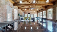 This expansive single-floor space is a masterfully converted industrial building that seamlessly blends historic character with modern amenities. The architecture is defined by dramatic vaulted ceilings supported by exposed heavy timber trusses and wooden decking, all set against original exposed brick walls that lend warmth and texture throughout. Rows of tall, arched multi-pane windows line the perimeter, flooding the interior with natural light and offering views of the surrounding greenery and neighboring brick structures.

The highly polished concrete floors reflect the abundant light, enhancing the open, airy atmosphere of the space. The main area is thoughtfully arranged into distinct functional zones: a comfortable lounge area features tufted sofas and contemporary accent chairs arranged around area rugs, with a wall-mounted flat-screen television and vivid graffiti murals adorning the brick walls nearby. A recreation zone includes a billiards table and ping pong table, along with a dartboard, catering to social and leisure activities.

A café-style seating area near the windows provides high-top tables and chartreuse chairs, ideal for casual gatherings. A compact kitchenette with white cabinetry, dark countertops, and a stainless steel refrigerator supports events and everyday use. Ceiling fans and pendant lighting complete this dynamic, character-rich space.