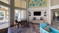 Located at River Park Apartments in Orlando, Florida, this single-level amenity facility is designed to support an active and social lifestyle. The centerpiece is a dramatic clubhouse lounge boasting soaring double-height ceilings, an octagonal tray ceiling detail, and bold teal pendant chandeliers. Walls of floor-to-ceiling arched windows and French doors flood the space with natural light and offer unobstructed views of the resort-style pool and outdoor seating area. Plush teal armchairs, patterned area rugs, and coordinated accent pieces give the lounge a polished, inviting ambiance. Dining tables positioned near the pool-facing glass doors provide a seamless indoor-outdoor connection. A small kitchenette area with wood cabinetry and a sink supports the entertaining space. The fitness center is well-appointed and energetically designed, featuring a vibrant motivational mural in teal, orange, and gold with words like "Believe," "Passion," and "Balance." It is equipped with a full row of treadmills with digital screens, ellipticals, stair climbers, and strength-training machines on durable rubber flooring. A dedicated racquetball court with a light hardwood floor and clearly marked red boundary lines rounds out the athletic offerings, providing residents with a versatile and comprehensive recreational facility.
