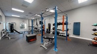 This fitness center is a comprehensively outfitted single-floor space designed to support a wide range of workout disciplines. The room is finished with neutral gray walls, a drop tile ceiling with recessed panel lighting, and durable speckled rubber flooring that provides both traction and impact absorption throughout the entire area.

At the heart of the space stands a prominent blue steel power rack equipped with pull-up bars, a barbell, suspension training straps, resistance bands, and a hanging heavy bag — making it a versatile hub for strength and functional training. An adjustable weight bench and a plyo box are positioned nearby for additional exercise variety.

Along one wall, a three-tier dumbbell rack holds a full set of free weights, accompanied by a wall-mounted flat-screen television. The cardio zone features multiple treadmills, an elliptical, a stationary bike, and additional cardio equipment, all positioned near windows that bring in natural light and outdoor views of surrounding greenery. A cable machine rounds out the strength training options.

The space also includes exercise mats, medicine balls, and battle ropes, supporting flexibility and high-intensity interval training. A wall clock and clearly defined zones keep the environment organized and functional. Overall, the fitness center offers a clean, professional ambiance suited for a broad spectrum of fitness routines.