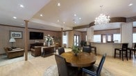 This single-floor space combines a dining area and living room in an open-plan layout, creating a seamless and spacious environment. The dining area is anchored by a rich wooden round table surrounded by dark leather chairs, positioned beneath a stunning crystal chandelier that adds a touch of glamour. Adjacent to it, the living area features a wall-mounted flat-screen TV, a console table adorned with floral arrangements, and leather seating, contributing to a warm and inviting atmosphere.

The interior is finished with warm neutral tones on the walls, complemented by crown molding and light hardwood flooring. Structural columns add architectural interest while subtly defining the different zones within the open space. Recessed lighting throughout provides even, ambient illumination, working in harmony with the chandelier.

Natural light filters into the space through multiple windows and glass-paneled doors, enhancing the bright and airy feel. A bar or counter area is visible toward the back, adding a functional element. Overall, the space balances elegant design details with a comfortable, welcoming ambiance suitable for both entertaining and relaxing.
