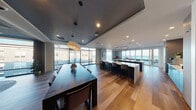 This sophisticated single-floor space showcases a seamlessly integrated open-plan layout designed for both functionality and social engagement. Wide-plank hardwood flooring flows throughout, unifying the distinct zones within the space.

The kitchen area is a standout feature, anchored by two large white quartz-topped islands with dark navy cabinetry beneath. A striking geometric diamond-patterned accent wall in light stone tones serves as a refined backdrop, while a curved linear pendant light adds a contemporary focal point overhead. The kitchen is well-equipped for hosting, with ample counter seating provided by sleek upholstered barstools.

Adjacent to the kitchen, a generous dining area accommodates a large dark wood communal table beneath an open-frame pendant light fixture, ideal for group gatherings. A wall-mounted television and curated artwork add to the functional yet stylish character of the dining zone.

The lounge area transitions naturally from the dining space, featuring a deep teal accent wall, ambient wall sconces, framed artwork, and plush seating arranged on a round area rug, creating an intimate and welcoming retreat. A bookshelf adds a touch of personality and warmth.

Floor-to-ceiling windows and glass doors on multiple sides bathe the entire space in natural light while framing expansive views of the surrounding waterfront and skyline.