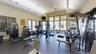 This single-floor fitness center offers a comprehensive workout environment designed to accommodate a variety of exercise routines. The space is generously sized, providing ample room for both cardio and strength training equipment without feeling crowded. Large windows line the walls, flooding the interior with natural light and offering views of the surrounding greenery, creating an open and energizing atmosphere. The light yellow walls and rubber-floored workout area contribute to a clean, functional aesthetic. A ceiling fan overhead aids in air circulation throughout the space. The cardio section features multiple treadmills and elliptical machines, while the strength training area includes a multi-station cable machine, weight benches, and a variety of resistance equipment. Wall-mounted mirrors and a flat-screen television add to the practicality and comfort of the space. Track lighting along the ceiling ensures the room remains well-lit during all hours. The layout is thoughtfully organized, allowing users to move freely between equipment. Overall, this fitness center combines functionality with a bright, welcoming ambiance suited for a thorough and enjoyable workout experience.