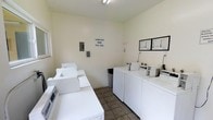 This shared laundry facility is a dedicated, single-floor space designed to serve residents with practicality and convenience in mind. The room features a row of top-loading washing machines along one side and a corresponding bank of dryers on the opposite side, providing ample capacity for simultaneous use. The machines are arranged in an organized, easy-to-navigate layout that maximizes the use of the available space. The interior is finished with neutral, light-colored walls and durable tile flooring, creating a clean and utilitarian atmosphere. Overhead lighting ensures the space is well-lit throughout. A posted sign on the wall clearly indicates the laundry room's operating hours, helping residents plan their laundry schedules accordingly. An informational board is mounted near the dryers, likely displaying usage instructions and facility guidelines. The room also includes a small utility sink and a waste bin for added convenience. A window allows for a degree of natural light to enter the space. Overall, this laundry facility is a well-maintained, functional communal amenity designed to meet the everyday needs of its users efficiently.