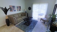 This single-floor residence offers a welcoming and functional layout designed for comfortable everyday living. The living area serves as the heart of the space, characterized by soft, neutral-toned carpeting that adds warmth underfoot and a clean, understated aesthetic throughout. Large sliding glass doors dominate one wall, allowing an abundance of natural light to flood the room while providing direct access to an outdoor area — a standout feature that enhances both the brightness and the sense of openness in the space. Vertical blinds offer flexible light control and privacy as needed. The living area is well-proportioned, offering enough room for a comfortable seating arrangement centered around a stylish coffee table, with wall space available for artwork and décor. The soft color palette of the walls complements the neutral flooring, creating a calm and cohesive atmosphere throughout. The overall ambiance of this residence is light, airy, and move-in ready, with a straightforward single-level layout that prioritizes ease of flow and livability. The combination of natural light, neutral finishes, and practical design elements makes this a versatile space suitable for a variety of lifestyles.