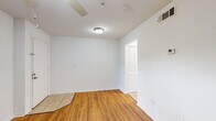 This single-story space in Gainesville, Florida offers a straightforward, functional layout with a clean, neutral aesthetic. The interior is finished with warm-toned hardwood-style flooring throughout the main living areas, transitioning to light tile in the kitchen and bathroom zones, creating a clear visual distinction between spaces.

The main living area is open and bright, featuring white walls, a ceiling fan with a light fixture, and smooth ceilings with centrally placed flush-mount lighting. The entry opens directly into this space, which connects to the kitchen via a wide doorway. Louvered bifold closet doors provide additional storage access visible from multiple vantage points.

The kitchen is equipped with white cabinetry, a white refrigerator, and a freestanding range, all set atop light tile flooring. A window brings natural light into the space, and the compact layout keeps everything within easy reach.

The bedroom features the same warm hardwood-style flooring, a ceiling fan, a window with blinds offering natural light, and a louvered bifold closet. The bathroom includes tile flooring, a white vanity with an undermount sink and stone-look countertop, a framed mirror, and brushed gold hardware accents, lending a subtle modern touch.