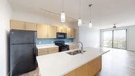 Located at 112 W Mishawaka Ave in Mishawaka, Indiana, this single-floor unit presents a contemporary, well-appointed interior with a clean, modern aesthetic. The open-concept kitchen serves as a central highlight, featuring light wood cabinetry, a large island with a white quartz-style countertop, a stainless steel double-basin sink with a pull-down faucet, and pendant lighting suspended above. Black appliances—including a refrigerator, electric range, and over-the-range microwave—complement the teal accent wall behind the cooking area. Track lighting along the ceiling provides additional illumination. Wood-look plank flooring runs consistently throughout the unit, lending warmth and visual continuity to each space. The living area is bright and open, benefiting from natural light through large windows fitted with horizontal blinds, as well as a sliding glass door with vertical blinds that opens to an outdoor view. Bedrooms are generously sized with neutral-toned walls and ample natural light. The bathrooms feature light wood vanities with white countertops, pendant lighting, and mirrored medicine cabinets. A dedicated walk-in closet with built-in shelving is accessible from the primary bathroom area, providing practical storage. Overall, the unit offers a cohesive, modern living environment.