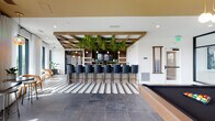 This expansive single-floor amenity space blends modern design with natural elements to create a dynamic, multi-functional environment. The open layout is anchored by a striking full-length kitchen featuring a substantial marble-topped island with bold veining, stainless steel cabinetry, wood countertops, and a vivid green ceramic tile backsplash extending to the ceiling. A professional-grade gas cooktop, range hood, and open shelving complete the well-equipped culinary area.

Overhead, a series of exposed wood beam lattices runs the length of the space, providing architectural definition while supporting cascading greenery and pendant lighting that adds warmth throughout. Flooring transitions between large-format stone tile and wood planks, delineating functional zones seamlessly.

A dedicated bar area features a row of sleek black leather barstools set beneath a dramatic overhead trellis draped in trailing plants, with a large-screen television and pendant globe lights creating an inviting social hub. A billiards table nearby adds a leisure element to the space.

Lounge seating areas offer comfortable sectional sofas beneath dramatic hanging botanical installations, positioned beside floor-to-ceiling windows that frame elevated city views. Intimate nook seating with dark walls and natural light provides quieter retreat spaces, while sheer-draped windows elsewhere flood the room with abundant natural light.