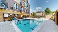 Located at 360 N First St in Minneapolis, Minnesota, this outdoor amenity courtyard is a well-appointed recreational space nestled within the grounds of a contemporary multi-story building. The centerpiece of the courtyard is a generously sized outdoor swimming pool with vivid blue tile accents, clearly marked depth indicators, and easy-access entry steps — ideal for both leisurely swims and relaxation. Adjacent to the pool is a hot tub, providing an additional amenity for residents. The pool deck is surrounded by ample concrete patio space lined with a variety of lounge seating options, including sun loungers, canopied recliners, and plush circular daybeds, offering residents multiple ways to enjoy the outdoors. A dedicated grilling station equipped with a full-sized grill and outdoor dining furniture provides a functional outdoor entertaining area. Decorative planters with seasonal flowers and lush greenery add warmth and color throughout the space. The courtyard is enclosed by a combination of stone masonry walls and metal fencing, offering both privacy and open views of the surrounding neighborhood and mature trees. The multi-story building rises above, with private balconies overlooking the courtyard, reinforcing the urban residential character of this vibrant North Loop Minneapolis location.