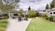 Located at 2949 Portage Bay W in Davis, California, this thoughtfully designed outdoor communal space offers a comprehensive range of amenities for relaxation and recreation. The layout is anchored by a spacious concrete patio that flows seamlessly between distinct activity zones.

At one end, a pergola-covered structure provides a shaded retreat equipped with ceiling fans, an outdoor television, and a dining table with wicker seating — ideal for al fresco gatherings. Adjacent to the covered area, a central fire pit surrounded by Adirondack chairs creates a welcoming focal point for casual socializing.

A well-maintained artificial turf lawn stretches alongside the patio, offering a clean, green surface suited for lawn games, including a cornhole set visible on the grounds. The surrounding landscape features layered plantings of ornamental grasses, manicured shrubs, and mature trees that provide privacy and a natural, park-like atmosphere.

The neighboring structures are modern in design, clad in gray board-and-batten siding and equipped with rooftop solar panels, reflecting an eco-conscious community ethos. Warm wood fencing defines the perimeter, adding visual warmth and enclosure. The overall ambiance is open, bright, and inviting, making this outdoor space a standout communal amenity.