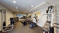 Located at 150 Chateau Terrace in Athens, Georgia, this fitness center offers a well-organized, single-floor workout facility designed to accommodate a comprehensive range of exercise routines. The space features durable rubber flooring throughout, providing both safety and comfort during workouts. Recessed lighting illuminates the room evenly, creating a bright and functional environment.

The cardio section is well-stocked with multiple SportsArt treadmills, elliptical trainers, and an upright stationary bike, offering ample options for aerobic exercise. A dedicated strength training area includes a multi-function cable machine and a weight bench, suitable for a variety of resistance exercises. A freestanding dumbbell rack lines one wall, providing a range of free weight options for targeted training.

A large wall-mounted flat-screen television is positioned prominently at one end of the room, keeping members entertained during their workouts. Wall mirrors expand the sense of space and allow users to monitor their form. The facility also includes a recycling bin and cleaning supplies, reflecting a commitment to cleanliness and sustainability. A windowed pass-through and an adjoining room add to the overall functionality of the space. Access is provided through both a standard and a French-style door.