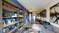 This single-floor space showcases a thoughtfully designed open-concept layout where the living, dining, and kitchen areas flow together naturally. A striking deep blue accent wall serves as a defining architectural feature, adding depth and character to the interior. A rustic, industrial-style wooden bookshelf with metal cross-bracing anchors the living area, offering both storage and display space while reinforcing the space's eclectic aesthetic.

The kitchen is outfitted with dark cabinetry, stone countertops, a tile backsplash, and stainless steel appliances, including a full-sized refrigerator. Pendant lighting fixtures hang above a compact dining area with a wooden table and modern bar-style stools, creating a cozy and functional dining nook.

The living area features warm wood-tone flooring throughout, complemented by layered area rugs and a neutral-toned sofa accented with blue decorative pillows. Track-style lighting and pendant fixtures provide warm, targeted illumination throughout the space. The overall ambiance balances industrial and contemporary design elements, creating a stylish, cohesive, and welcoming environment.