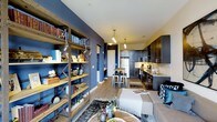 This single-floor space showcases a thoughtfully designed open-concept layout where the living, dining, and kitchen areas flow together naturally. A striking deep blue accent wall serves as a defining architectural feature, adding depth and character to the interior. A rustic, industrial-style wooden bookshelf with metal cross-bracing anchors the living area, offering both storage and display space while reinforcing the space's eclectic aesthetic.

The kitchen is outfitted with dark cabinetry, stone countertops, a tile backsplash, and stainless steel appliances, including a full-sized refrigerator. Pendant lighting fixtures hang above a compact dining area with a wooden table and modern bar-style stools, creating a cozy and functional dining nook.

The living area features warm wood-tone flooring throughout, complemented by layered area rugs and a neutral-toned sofa accented with blue decorative pillows. Track-style lighting and pendant fixtures provide warm, targeted illumination throughout the space. The overall ambiance balances industrial and contemporary design elements, creating a stylish, cohesive, and welcoming environment.