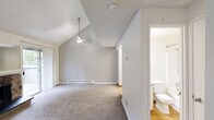 Located in University Place, WA, this two-floor home offers a thoughtfully laid-out living space spread across two levels. The main floor centers around a spacious living room highlighted by soaring vaulted ceilings and a ceiling fan, creating an airy, open atmosphere. A striking stone fireplace serves as a focal point, while a sliding glass door floods the room with natural light and provides access to an outdoor area with views of surrounding greenery. The kitchen is accessible directly from the living area and features white cabinetry and appliances with hardwood-style flooring. Baseboard heating is present throughout, ensuring comfort year-round. A staircase leads to the upper level, where a loft space sits beneath a sloped roofline, offering a bright and versatile area thanks to a wide horizontal window overlooking the natural surroundings. The bathroom on the main floor is clean and functional, featuring a full tub and shower combination with a handheld showerhead and classic chrome fixtures. A dedicated utility room rounds out the layout, adding practical storage and laundry capabilities. The overall aesthetic is neutral and understated, with consistent carpet flooring on both levels and a light color palette throughout.
