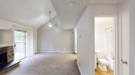 Located in University Place, WA, this two-floor home offers a thoughtfully laid-out living space spread across two levels. The main floor centers around a spacious living room highlighted by soaring vaulted ceilings and a ceiling fan, creating an airy, open atmosphere. A striking stone fireplace serves as a focal point, while a sliding glass door floods the room with natural light and provides access to an outdoor area with views of surrounding greenery. The kitchen is accessible directly from the living area and features white cabinetry and appliances with hardwood-style flooring. Baseboard heating is present throughout, ensuring comfort year-round. A staircase leads to the upper level, where a loft space sits beneath a sloped roofline, offering a bright and versatile area thanks to a wide horizontal window overlooking the natural surroundings. The bathroom on the main floor is clean and functional, featuring a full tub and shower combination with a handheld showerhead and classic chrome fixtures. A dedicated utility room rounds out the layout, adding practical storage and laundry capabilities. The overall aesthetic is neutral and understated, with consistent carpet flooring on both levels and a light color palette throughout.