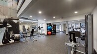 This fitness center is a comprehensive, thoughtfully designed workout facility spread across a single level, offering a broad array of equipment to support diverse training needs. The main exercise area features durable speckled rubber flooring throughout, providing both comfort and safety during workouts. Recessed lighting illuminates the space evenly, while multiple windows along the walls allow natural light to filter in and provide views of the exterior surroundings.

The cardio zone is well-appointed with treadmills, upright bikes, and a rowing machine, positioned near the windows to make workouts more engaging. The strength training section includes a variety of cable machines, a functional trainer, a leg press, a dumbbell rack, and a vertical knee raise station. A dedicated storage rack holds an assortment of medicine balls, stability balls, and kettlebells, supporting functional and dynamic training routines.

Floor-to-ceiling mirrors are strategically placed throughout, visually expanding the space and allowing users to monitor their form. A staircase with a modern metal railing is visible within the space, adding architectural dimension. A standout feature is the large decorative mural referencing the Old Chisholm Cattle Trail, incorporating a bold longhorn graphic and a historical map, lending the facility a distinctive regional character and visual focal point.
