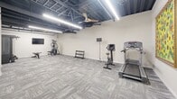 Located at 2500 Kelly Rd in Warrington, PA, this dedicated exercise room offers a well-organized and functional fitness space designed to support a variety of workout routines. The room features an open, single-floor layout with a generous footprint, providing ample space to move freely between equipment stations.

The interior is finished with durable, patterned carpet tiles in neutral grey tones, offering both comfort and practicality underfoot. Crisp white walls contrast with the exposed industrial-style ceiling, which showcases dark-painted structural elements, overhead ductwork, and bright linear LED lighting that ensures the space is well-lit throughout. Ceiling fans contribute to air circulation, enhancing comfort during workouts.

The exercise room is equipped with a variety of fitness machines, including a treadmill, upright stationary bike, and a multi-function strength training station, along with a three-tier dumbbell rack. Multiple wall-mounted flat-screen monitors are positioned throughout the room, allowing users to stay entertained or track their workouts. A built-in cubby storage unit provides convenient space for personal belongings. Colorful wall art adds a welcoming touch near the entry. The layout is thoughtfully arranged to maximize usability while maintaining an open, uncluttered environment.
