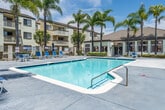 Located at 11902 Central Ave in Chino, California, this outdoor amenity space serves as the communal recreational hub of a multi-story residential complex. The centerpiece of the area is a generously sized swimming pool with clear, blue-tinted water, offering ample space for both leisure and lap swimming. The pool deck is finished in a smooth concrete surface and is lined with blue lounge chairs, providing residents with comfortable outdoor seating ideal for relaxing under the Southern California sun. Adjacent to the main pool is a separate spa, enclosed by a wrought iron security gate and tiled in classic blue, offering a more intimate setting for relaxation. Towering palm trees are strategically positioned throughout the space, adding a tropical ambiance and providing natural shade along the perimeter. The surrounding landscape includes manicured hedges and ornamental trees that enhance the overall aesthetic. A dedicated clubhouse or community building is visible poolside, featuring large windows and a covered patio area, suggesting additional indoor amenities. The multi-story residential buildings that frame the space feature balconies overlooking the pool area. The entire recreational zone is enclosed by a secure fencing system, ensuring privacy and safety for residents throughout the community.