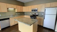 This single-floor space is thoughtfully designed to offer both functionality and style. The kitchen serves as a standout feature, showcasing light maple cabinetry that extends to the upper walls, providing generous storage throughout. Granite countertops with warm, earthy tones run along the full perimeter and are echoed on the central kitchen island, which offers additional prep space and serves as a natural focal point of the room. Stainless steel appliances, including a range with electric cooktop, an over-the-range microwave, and a refrigerator, complement the modern aesthetic. Rich, dark hardwood flooring anchors the space and provides a striking contrast to the lighter cabinetry and countertop surfaces. Sage-toned walls add a subtle warmth to the overall palette, tying together the natural textures found throughout the kitchen. The layout is practical and open, allowing for ease of movement and an intuitive workflow. The elevated cabinetry reaching toward the ceiling maximizes vertical storage, making the space as efficient as it is visually appealing. Overall, the kitchen exudes a clean, contemporary character well-suited for everyday living and entertaining alike.