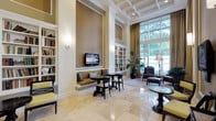 This sophisticated common lounge and library area exudes a refined, upscale ambiance, thoughtfully designed to offer both comfort and functionality. The space is anchored by dramatically soaring ceilings adorned with a coffered detail, creating an immediate sense of grandeur upon entry. Polished travertine tile flooring runs throughout, lending a clean, luminous quality to the interior.

Generous floor-to-ceiling windows line one side of the room, framed by flowing gold drapery and allowing an abundance of natural light to pour in while offering views of lush greenery outside. Multiple distinct seating zones are arranged throughout the space, including a built-in upholstered bench nook with a wall-mounted television, intimate bistro-style tables with dark wood chairs, and a more relaxed lounge area with rich leather armchairs and ottomans.

Flanking the main area are tall, built-in white bookshelves filled with an extensive collection of books, serving as both a functional library and a striking architectural feature. Warm, textured grasscloth-style wall treatments in neutral tones add depth and warmth to the interior. Potted tropical plants are placed throughout, introducing organic vitality and softening the formal elegance of the design. A corridor lined with structural columns connects to additional areas, completing this well-appointed, inviting retreat.