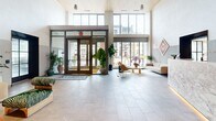 Located in Washington, DC's vibrant NoMa neighborhood, the lobby of The Florian at 1 Florida Ave NE makes an immediate impression with its dramatic double-height ceilings and an abundance of natural light streaming through expansive floor-to-ceiling windows flanking the main entrance. Large-format stone tile flooring extends throughout, providing a clean, polished foundation that complements the warm, light wood paneling that clads the soaring interior walls.

At the heart of the space sits a commanding reception desk finished in richly veined marble, accented by warm underlighting that adds depth and elegance. The seating lounge invites visitors to linger, furnished with contemporary sofas, accent chairs, and a curated selection of indoor plants that bring a natural, refreshing energy to the interior. A grand piano tucked into an alcove alongside original artwork adds a cultural and refined touch.

A wide, well-appointed elevator corridor connects the main lobby to the rest of the building, lined with leather bench seating, light wood wall panels, dark-framed elevator doors, and The Florian branded floor mats. The interplay of natural materials, warm tones, and modern design details creates a cohesive, welcoming ambiance that reflects the building's elevated character throughout this generously scaled ground-floor space.