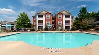 Located at 916 Sugar Gap Rd in Wake Forest, North Carolina, this community amenity space serves as a central gathering hub within a well-appointed residential community. The single-floor structure is designed with a classic exterior, featuring warm taupe lap siding, crisp white trim, and stone-accented columns that lend a polished, welcoming character to the entrance.

A generously sized covered porch extends from the building, offering an inviting outdoor seating area furnished with comfortable lounge chairs and colorful accent rugs — an ideal spot to relax and take in views of the surrounding greenery and pool area. Large windows with white shutters allow abundant natural light to fill the interior while maintaining privacy.

The centerpiece of the outdoor amenity space is a large, sparkling resort-style swimming pool, framed by a spacious sun deck lined with lounge chairs and shaded by bright teal umbrellas. The pool's clear blue water and well-maintained deck create a vibrant, vacation-like atmosphere. The surrounding landscape is meticulously maintained, with manicured shrubs, colorful flowering plants, and lush green trees providing a serene natural backdrop.

The broader community setting features attractive multi-story residential buildings with red and neutral-toned exteriors, private balconies, and stone detailing, all contributing to a cohesive and upscale aesthetic throughout the property.