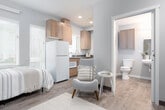 This single-floor studio unit offers a smart, open-concept layout that efficiently combines living, sleeping, and cooking areas into one cohesive space. The main room features warm wood-tone flooring throughout, soft neutral wall tones in muted blues and grays, and a wall-mounted climate control unit, creating a comfortable and contemporary ambiance. Large windows fitted with white horizontal blinds allow for generous natural light, brightening the sleeping and living area. A dedicated kitchenette lines one wall and is outfitted with warm wood cabinetry extending to the ceiling for ample storage, a full-size refrigerator, a compact microwave, a sink with dark countertop, and an additional window that enhances ventilation and light. The sleeping area accommodates a full-size bed and benefits from a closet with a full-panel door, providing practical storage. The private bathroom is cleanly appointed with a wall-mounted sink, toilet, overhead wood cabinetry, a mirrored medicine cabinet, and a shower with a curtain enclosure, all finished in coordinating neutral tones. Durable wood-look flooring runs consistently through the bathroom as well, tying the unit together with a unified, modern design sensibility.