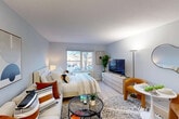 Located at 1850 Post Rd in Warwick, Rhode Island, this efficiently designed studio offers a smart, open-concept layout that seamlessly integrates the living, sleeping, dining, and kitchen areas into a single cohesive space. Light wood-tone flooring runs throughout, lending warmth and visual continuity to the interior.

The main living area is bright and welcoming, with a large window that allows natural light to fill the room and provides outdoor views of the surrounding landscape. Soft, neutral wall tones in a cool gray-blue palette complement the contemporary aesthetic. The space comfortably accommodates a full sleeping area alongside a dedicated living zone, complete with a sofa and glass coffee table arrangement anchored by a patterned area rug.

The kitchenette is thoughtfully appointed with gray wood-grain cabinetry, a stainless steel refrigerator, a microwave, and a single-basin sink — delivering essential functionality in a compact footprint. A small dining area with seating sits adjacent to the kitchen, making the most of the open floor plan. An open shelving unit near the entrance provides additional storage and display space.

The bathroom features tile flooring, a granite-topped vanity, a full bathtub with shower, and clean, understated finishes. The entrance provides a natural transition point into the living space, keeping the layout organized and functional.