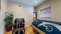 This single-floor wellness space is thoughtfully designed to promote relaxation and recovery through dedicated therapeutic amenities. The treatment room serves as the heart of the suite, housing a large float therapy pod finished in a bold blue and white exterior — a state-of-the-art sensory deprivation vessel intended for deep relaxation and mental clarity. Positioned alongside it is a full-body zero-gravity massage chair, offering an additional layer of restorative comfort before or after a float session. Warm-toned hardwood flooring runs throughout, lending the space a clean and inviting aesthetic. Natural light filters in through slatted window blinds, contributing to a soft, controlled ambiance well-suited for therapeutic use. Zen-inspired wall art featuring bamboo, stones, and nature motifs reinforces the calming atmosphere, while lush potted plants add organic texture and a sense of tranquility. The neutral, light-gray walls keep the environment visually serene and uncluttered. Interior doorways connect the treatment area to adjacent spaces, and the overall layout flows smoothly to support a seamless client experience. The space is compact yet purposeful, with every element carefully chosen to foster a sense of calm and well-being.