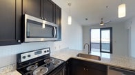 This well-appointed single-floor space showcases a thoughtfully designed kitchen that serves as a central highlight. The kitchen features rich, dark cabinetry with sleek hardware, offering a striking contrast against the light granite countertops. A classic white subway tile backsplash adds a timeless touch, while stainless steel appliances — including a range and over-the-range microwave — provide a polished, modern aesthetic. The undermount stainless steel sink is positioned at a peninsula, enhancing both functionality and flow. A pendant light fixture hangs overhead, casting focused illumination over the workspace and complementing the ceiling fan visible in the adjacent living area. The open layout creates a seamless connection between the kitchen and the living space, which benefits from abundant natural light streaming through large sliding glass doors. These doors open directly to an outdoor deck or patio area, extending the living space and providing views of the surrounding environment. The crisp white walls and clean lines throughout contribute to a bright, contemporary ambiance that feels open and inviting. The overall design balances practicality with modern style, making it a functional and attractive space.