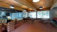 This single-floor amenity space is designed for leisure and social gathering, offering a well-appointed environment with a thoughtful blend of rustic and functional elements. The centerpiece of the room is a full-sized billiards table, positioned beneath a striking wood-planked ceiling supported by exposed wooden beams that lend the space a warm, lodge-inspired character. Pendant light fixtures with dark drum shades hang overhead, providing focused illumination throughout. Large windows line multiple walls, flooding the interior with natural light and offering views of the surrounding outdoor greenery and brick exterior. The patterned carpeted flooring in warm earth tones adds texture and comfort underfoot. A relaxed lounge area with upholstered armchairs is positioned to one side, ideal for casual conversation or spectating. On the opposite end, a dining area with round tables and cushioned chairs provides space for group gatherings or casual meals. The overall ambiance is inviting and versatile, combining recreational, social, and relaxation functions within a single cohesive space that feels both comfortable and well-utilized.