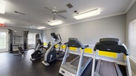 This fitness center is a thoughtfully designed, single-floor workout facility that offers a comprehensive range of equipment and a welcoming atmosphere for exercise. The space is finished in a crisp grey color palette, accented by bold yellow wall panels that add a dynamic, energetic character to the room. Crown molding and white wainscoting detail the walls, lending a polished, finished look uncommon in typical gym spaces.

The facility is well stocked with Matrix-brand cardio equipment, including multiple treadmills and elliptical trainers, as well as a stationary bike. A multi-station cable and strength machine anchors one end of the room, complemented by adjustable workout benches and free-weight accessories for resistance training. Dark rubberized flooring covers the entire space, providing both durability and safety underfoot.

A full-length mirror runs along one wall, creating an open, expansive feel while allowing users to monitor their form during workouts. Multiple ceiling fans and bright flush-mounted panel lights ensure the space remains comfortable and well-lit throughout the day. Natural light filters in through several windows with white blinds, and entry is provided via multiple doors. Framed artwork adorns the walls, adding a welcoming touch to this functional, well-maintained fitness space.