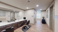 Located at 3640 Harmony Commons Drive in Spring, TX, this salon and wellness space is thoughtfully designed to provide a comfortable and functional environment for personal care services. The main salon area features two professional styling stations equipped with sleek, black hydraulic chairs positioned along an extended countertop with dark espresso cabinetry and a large wall mirror that enhances the sense of space. Recessed lighting throughout the space provides consistent, flattering illumination, while light wood-look flooring adds warmth and a clean aesthetic. A dedicated shampoo alcove is set apart with its own sink, cabinetry, and a large window fitted with blinds that allows natural light to filter in. Adjacent to the main area, a separate massage room offers a private, tranquil setting furnished with a full-length massage table and accented by calming landscape artwork. A small foyer connects the rooms, complete with a tufted accent bench and framed wall art, lending an inviting, polished feel. Glass-paneled entry doors contribute to the open and professional character of the space, making it well-suited for a senior living community or similar facility seeking on-site wellness amenities.