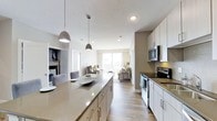 This single-floor space showcases a thoughtfully designed open-concept layout that integrates the kitchen, dining, and living areas into one cohesive environment. The kitchen is well-appointed with white cabinetry, dark countertops, and stainless steel appliances, including a range and microwave. A large central island with a light quartz surface serves as both a functional workspace and a casual dining area, anchored by stylish pendant lights overhead. The backsplash adds a subtle decorative touch behind the cooking area. Light wood-style flooring flows continuously throughout the space, reinforcing a clean and modern aesthetic. The open layout leads naturally into the dining and living areas, which benefit from an abundance of natural light streaming in through a large window or sliding glass door at the far end. The living area is spacious and inviting, with neutral tones that create a calm, welcoming ambiance. A laundry area with stacked or side-by-side units is visible, adding practical convenience. Overall, the space balances functionality with contemporary design.