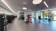 Located at 1042–1044 NW Seventh Street in Miami, this single-floor fitness center offers a thoughtfully designed workout environment that balances functionality with a polished, contemporary aesthetic. The space is organized into several distinct training zones, each equipped to support a comprehensive range of exercise routines.

The cardio area lines the perimeter wall of floor-to-ceiling windows, where treadmills, stationary bikes, and rowing machines are positioned to take advantage of lush outdoor views of surrounding greenery. Large pendant lights hang throughout, complementing the warm cove lighting that traces the ceiling edges and contributes to an inviting ambiance.

A dedicated strength-training zone houses a selection of cable machines, selectorized equipment, and multi-functional cable systems, all arranged with ample floor space between stations. Full-length mirrors along the walls visually expand the area and allow users to monitor form during workouts. A separate free weights section includes a neatly organized dumbbell rack and adjustable benches.

The entry corridor features glass-paneled doors, a wall-mounted water fountain, and smooth rubberized flooring that extends throughout the facility. A large video display screen serves as a focal point in one section of the space. Overall, the fitness center presents a clean, professional environment well-suited for diverse training needs.