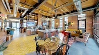 This expansive single-floor communal amenity space blends rustic lodge aesthetics with modern industrial design in a bold and energetic setting. Soaring ceilings clad in warm wood planks are supported by exposed structural beams and paired with visible ductwork, creating an open, loft-style atmosphere throughout. Large glass garage doors and floor-to-ceiling windows flood the interior with natural light while offering views of the surrounding outdoor areas.

The fully equipped communal kitchen serves as a central hub, anchored by a generous island with a marble-style countertop, bar-height seating with red stools, a professional-grade range with a copper hood, and ample cabinetry. Decorative chandeliers hang overhead, adding warmth above the vibrant, patterned tile floors in shades of teal, red, and yellow.

Adjacent dining and lounge areas feature eclectic furnishings, including patterned upholstered chairs, natural wood accent tables, and wall-mounted décor such as antler mounts and curated artwork. The game room showcases a full-size pool table beneath pendant lighting, flanked by a striking laser-cut black metal feature wall, a foosball table, and cozy lounge seating. Cedar-paneled walls, decorative bookshelves stocked with curated items, and thoughtful greenery complete the inviting, cabin-inspired character of this dynamic social space.